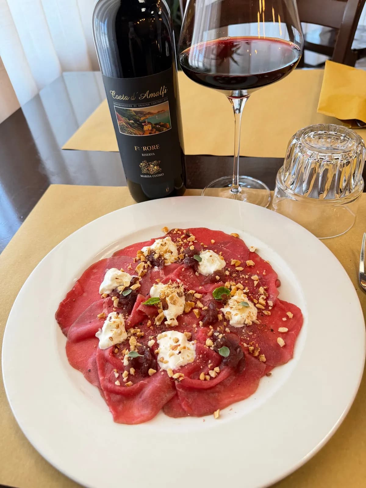 Un piatto di carpaccio su una tovaglietta gialla, con una bottiglia e un calice di rosso
